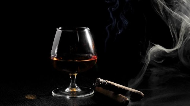 Luxury brandy. Glass with cognac, two cigars and smoke from cigar rotate on a black table against black background. Brandy, cognac, snifter, binge. Slow motion.