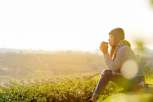 Asian Lifestyle Women Wearing Sweater, Sitting And Drinking Hot Coffee Or Tea Relax Outdoor In The Sunrise Morning Sunny Day At Tea Plantation Nature.  Lifestyle Concept.