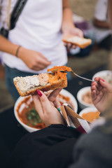 Woman hands holds chickpea hummus on spoon. The woman puts the hummus on a slice of bread. Outside party. Another person and bowls of hummus in the background. Close up