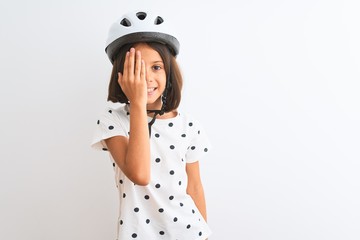 Beautiful child girl wearing security bike helmet standing over isolated white background covering one eye with hand, confident smile on face and surprise emotion.