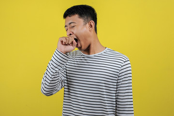 Portrait of funny Asian man yawning covering open mouth and showing a sleepy gesture. feeling being tired from hard work, isolated on yellow