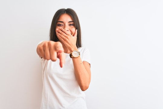 Young beautiful woman wearing casual t-shirt standing over isolated white background laughing at you, pointing finger to the camera with hand over mouth, shame expression