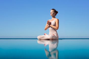 Side view of a beautiful young slender woman sitting in lotus position and meditating on the edge of the pool on a blue sky. Concept of yoga relaxation and meditation. Copyspace