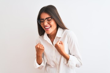 Young beautiful businesswoman wearing glasses standing over isolated white background very happy and excited doing winner gesture with arms raised, smiling and screaming for success