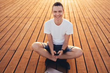 Portrait of a happy young male freelancer working on the street near the sea on a wooden platform with a laptop and smartphone on a sunny summer day. Remote work concept