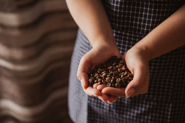 Woman's hands holding coffee beans. The woman is wearing a navy pinafore. Brown background.