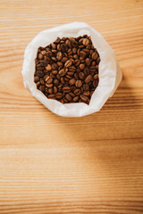 Roasted, brown coffee beans in white paper bag. Wooden background.