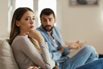 Obraz premium Worried young woman sitting on sofa at home and ignoring her boyfriend who is sitting next to her