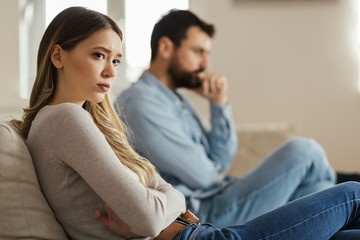 Worried young woman sitting on sofa at home and ignoring her boyfriend who is sitting next to her - obrazy, fototapety, plakaty