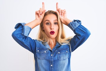 Young beautiful woman wearing casual denim shirt standing over isolated white background doing funny gesture with finger over head as bull horns