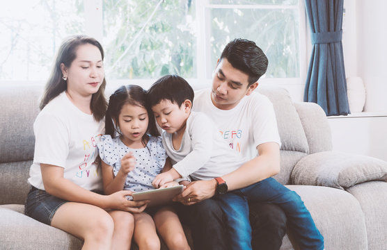 Young Parents Sitting On The Couch With Their Daughter And Son Using A Tablet In Living Room At Home. Asain Family Concept