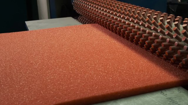 In production workshop for processing synthetic materials, orthopedic mattress production machine rotates with twisted rolls, pulls red sheet of paralon into its mechanism, desired relief of paralon