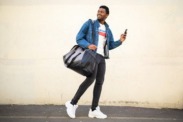 Full body happy young african american man walking with bag and mobile phone on street