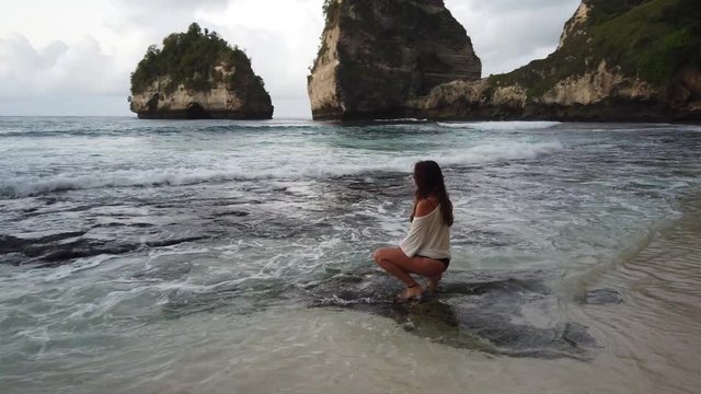 A young, brunette woman with long hair, dressed in a loose fit, oversized t-shirt, sitting by herself on a rock on the shore, while waves are splashing around her. Crouching down.