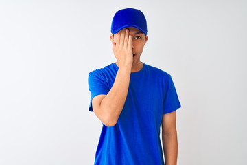Chinese deliveryman wearing blue t-shirt and cap standing over isolated white background covering one eye with hand, confident smile on face and surprise emotion.
