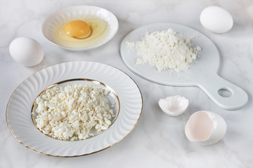 Ingredients for baking: cottage cheese, eggs, flour on white marble background.
