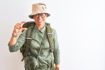 Middle age hiker woman wearing backpack canteen hat glasses over isolated white background smiling and confident gesturing with hand doing small size sign with fingers looking and the camera