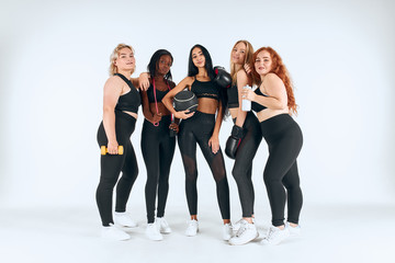 Fototapeta na wymiar Five smiling multiethnic women wearing sportive leggins and topics holding fitness ball, boxing gloves and other things isolated over white background