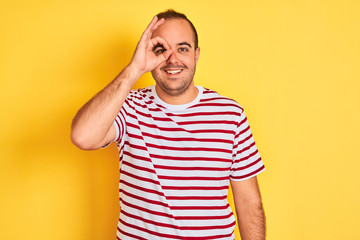 Young man wearing casual striped t-shirt standing over isolated yellow background doing ok gesture with hand smiling, eye looking through fingers with happy face.