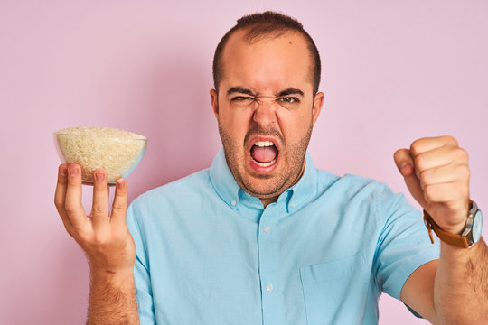 Young man holding bowl with rice standing over isolated pink background annoyed and frustrated shouting with anger, crazy and yelling with raised hand, anger concept