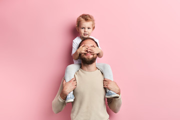 little boy closing his dad's eyes with palms, close up portrait, isolated pink background, studio...