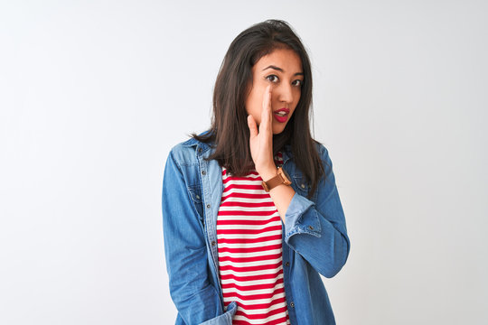 Young chinese woman wearing striped t-shirt and denim shirt over isolated white background hand on mouth telling secret rumor, whispering malicious talk conversation