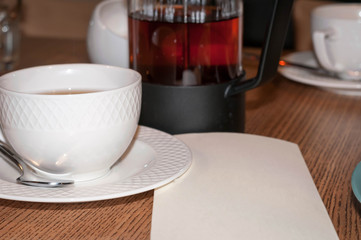 side view of white cup, glass kettle with dark tea and empty paper blank with copy space on wooden...