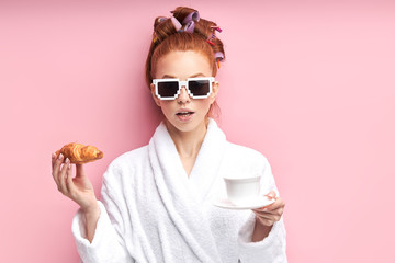 Caucasian girl after shower enjoying cup of tea with croissant. Isolated over pink background