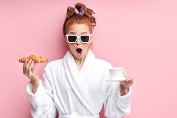 Young caucasian girl in white bathrobe stand with croissant and cup of tea. After shower in...