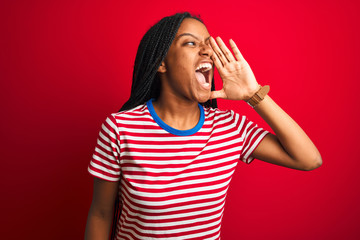 Young african american woman wearing striped t-shirt standing over isolated red background shouting and screaming loud to side with hand on mouth. Communication concept.
