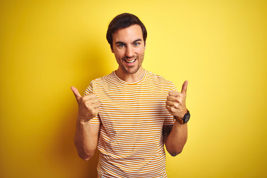 Young handsome man with tattoo wearing striped t-shirt over isolated yellow background success sign doing positive gesture with hand, thumbs up smiling and happy. Cheerful expression