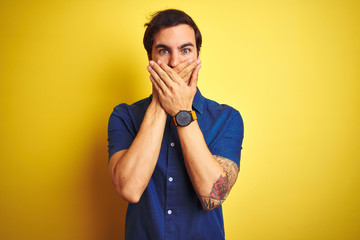 Young handsome man with tattoo wearing casual shirt standing over isolated yellow background shocked covering mouth with hands for mistake. Secret concept.