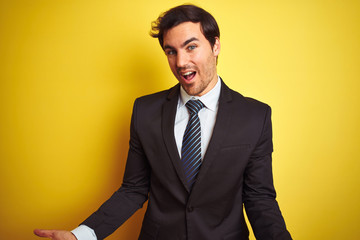 Young handsome businessman wearing suit and tie standing over isolated yellow background smiling cheerful with open arms as friendly welcome, positive and confident greetings