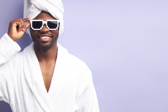 Young Man In White Bathrobe And Towel Look At Camera Smiling. Studio Portrait Against Purple Background