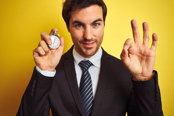 Fototapeta premium Young handsome businessman holding stopwatch standing over isolated yellow background doing ok sign with fingers, excellent symbol