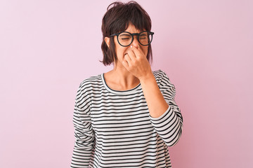 Young beautiful woman wearing striped t-shirt and glasses over isolated pink background smelling...