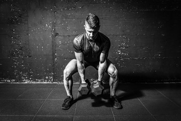Fototapeta na wymiar Young strong fit muscular sweaty man with big muscles strength cross workout training with dumbbells weights in the gym dark image with shadows real people black and white