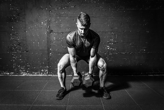 Young strong fit muscular sweaty man with big muscles strength cross workout training with dumbbells weights in the gym dark image with shadows real people black and white