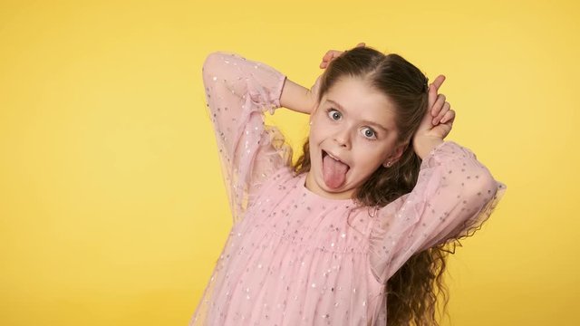 Portrait of cute little girl playfully making faces looking at camera. Caucasian kid girl in pink dress doing silly funny face at yellow background. Positive emotion. Medium shot in 4k, UHD