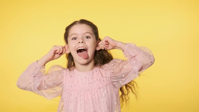 Caucasian kid girl in pink dress doing silly funny face at yellow background. Portrait of cute little girl playfully making faces looking at camera. Positive emotion. Medium shot in 4k, UHD