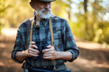 man with grey beard holding binoculars during hunting in autumn forest, looking away. Road background