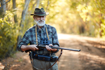 Older hunter man with grey beard holding gun to hunt on birds, ready to shoot. Forest background