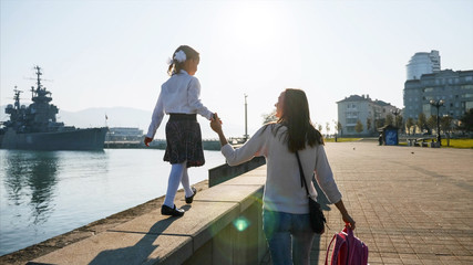 A little girl in school uniform with mother is walking on the railing of the promenade along the...
