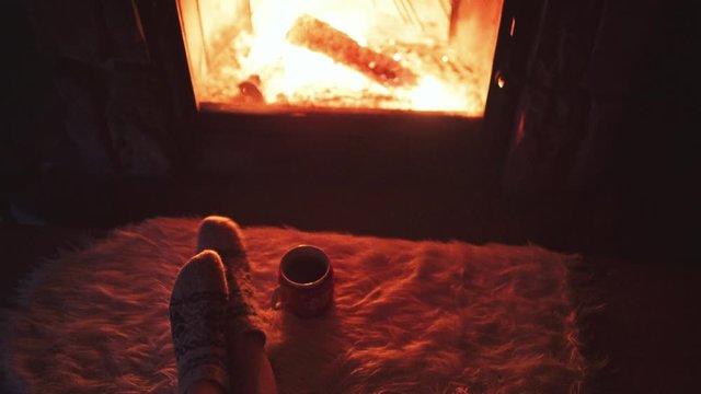 Feet in woollen socks by Burning Christmas Fireplace. 4K STABILIZED shot. Woman relaxes by warm fire with a cup of hot drink and warming up her feet. Close up. Winter and Christmas holidays concept.