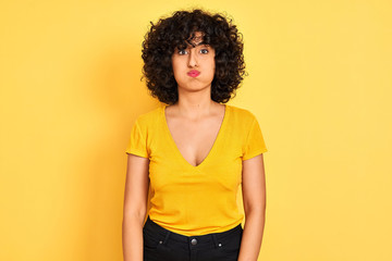 Young arab woman with curly hair wearing t-shirt standing over isolated yellow background puffing cheeks with funny face. Mouth inflated with air, crazy expression.
