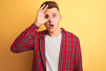 Young handsome man wearing casual shirt standing over isolated yellow background doing ok gesture shocked with surprised face, eye looking through fingers. Unbelieving expression.