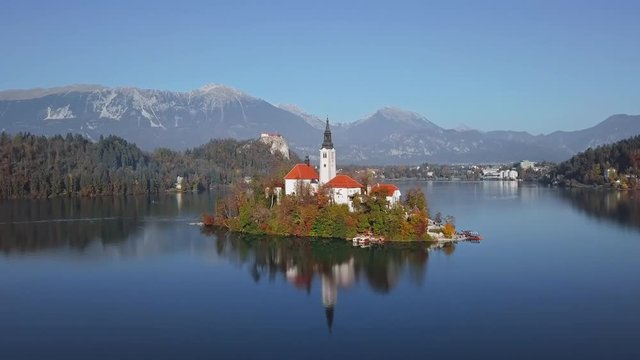 Flight around of Bled Island (Blejski otok) with pilgrimage church of the Assumption of Mary and Bled castle on Lake Bled over colorful autumn forest at clear sunny day, Slovenia