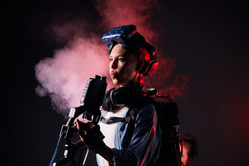 Beautiful girl playing virtual game, stand blowing the barrel of vr weapon, isolated over smoky...