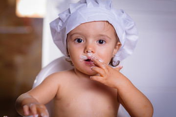 A sweet small chef cook in a white culinary cap the eating flour from fingers. Happy girl bullies...