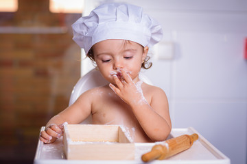 A sweet small chef cook in a white culinary cap the eating flour from fingers. Happy girl bullies...